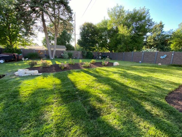 A sunny backyard featuring a lush green lawn, a landscaped flower bed with small bushes, and a tall fence in the back.
