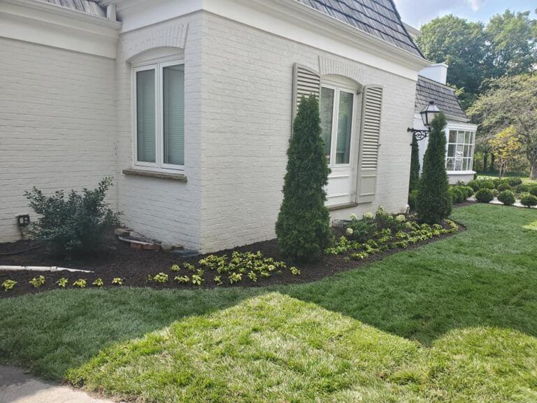 A cream-colored brick house features arched windows, shutters, and a garden bed planted with evergreen shrubs and groundcover.