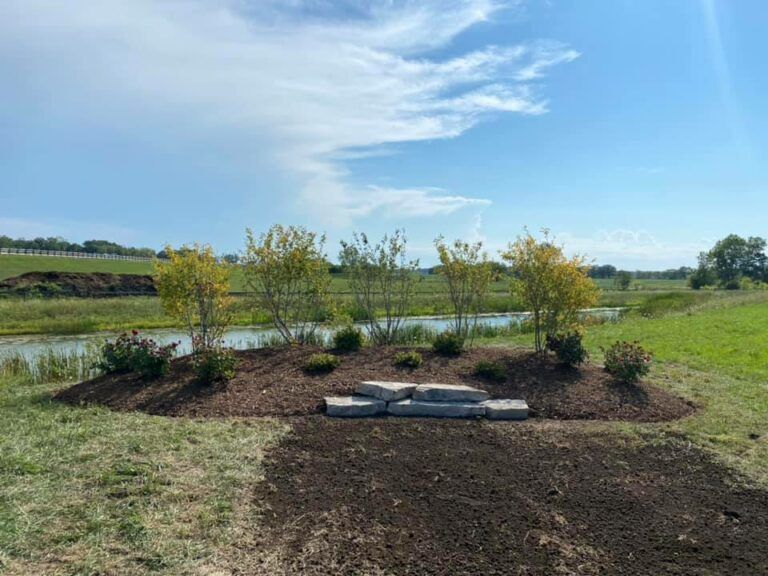 A landscaped mound with shrubs and layered stone steps sits beside a pond under a blue sky with thin, wispy clouds.