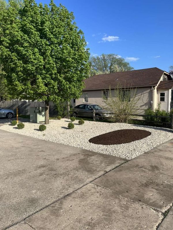 A landscape bed with white gravel, a large tree, small shrubs, and an oval mulch patch in front of a house.