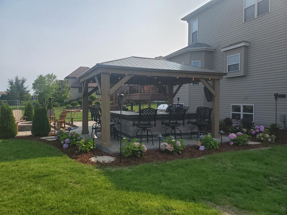 A backyard patio features a wooden gazebo covering an outdoor kitchen with bar seating, surrounded by landscaped bushes.