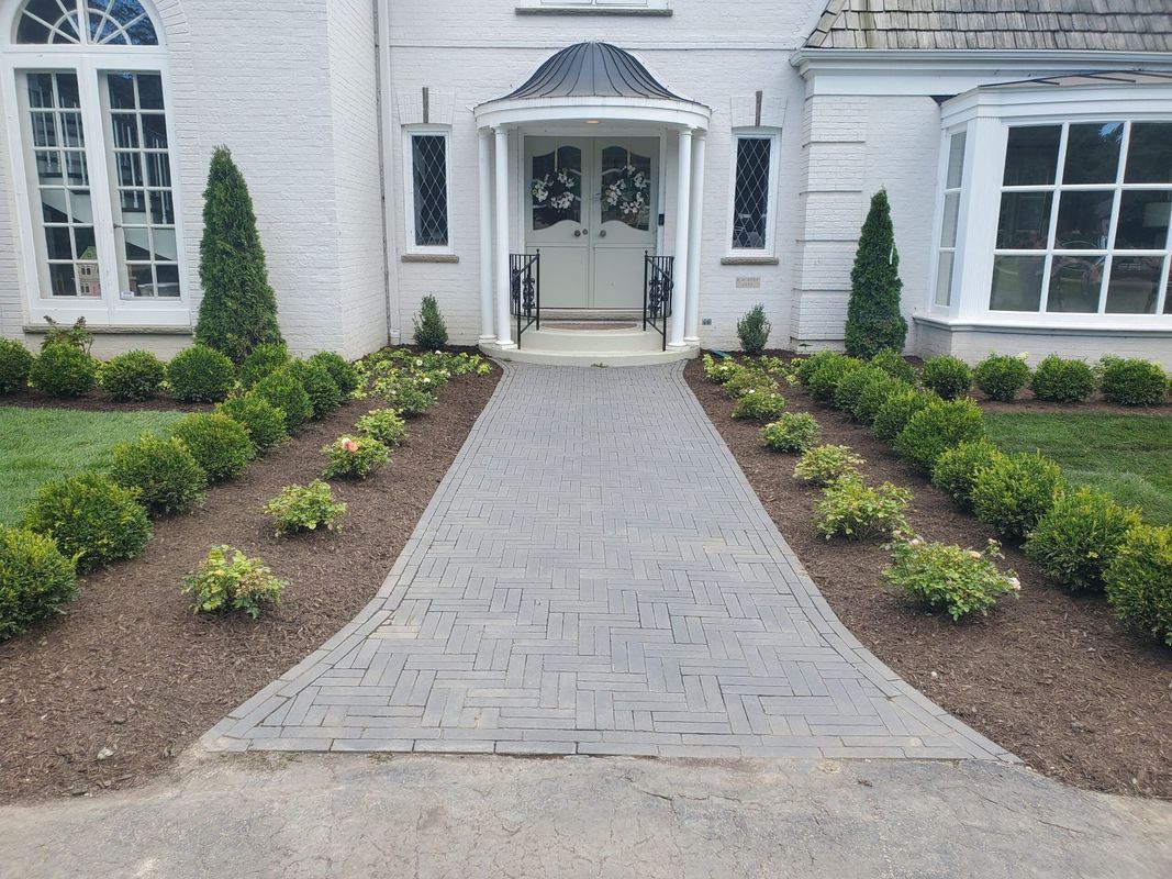 A gray patterned stone walkway leads to the white double-door entrance of a house, flanked by manicured green hedges.