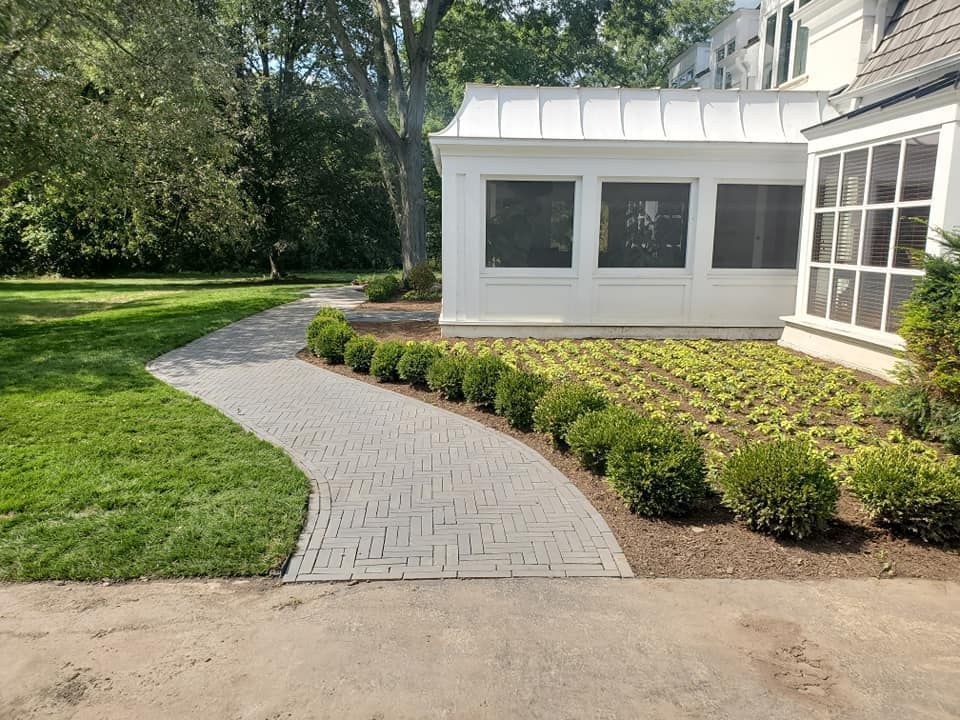 A gray stone pathway curves alongside a manicured row of small round bushes and a white house with large windows.