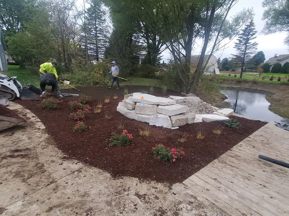Two workers landscape a garden bed with mulch and large stone blocks next to a pond and a paved walkway.