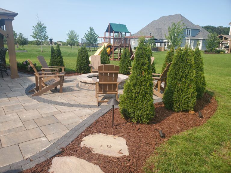 A backyard patio features a circular fire pit surrounded by wooden Adirondack chairs, evergreen shrubs, and stone pavers.