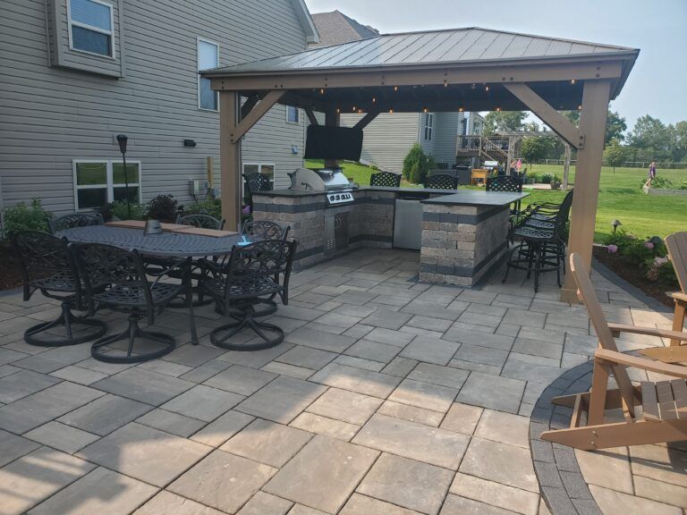 A stone patio featuring a covered outdoor kitchen, dining table with chairs, and a bar area with stools in a backyard.
