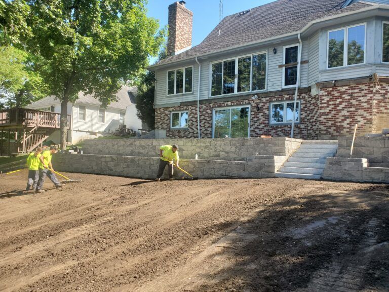 Three workers in high-visibility vests landscaping the front yard of a two-story home with a stone retaining wall and stairs.