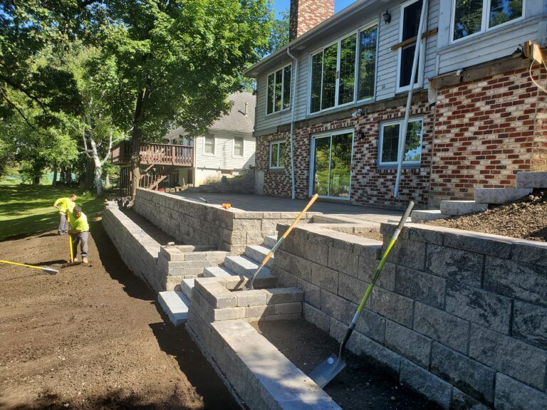 Workers prepare the soil in a yard next to a newly constructed multi-tiered stone retaining wall leading to a house.