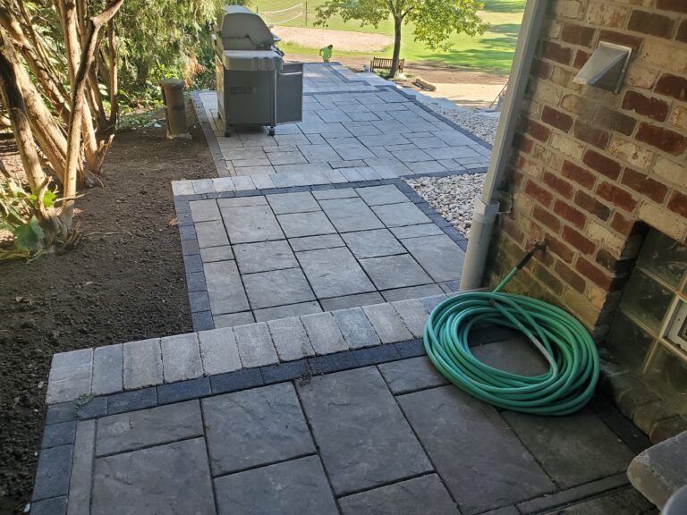 A multi-level stone patio with a grill, gray pavers, a dark border, and a green garden hose coiled by a brick wall.