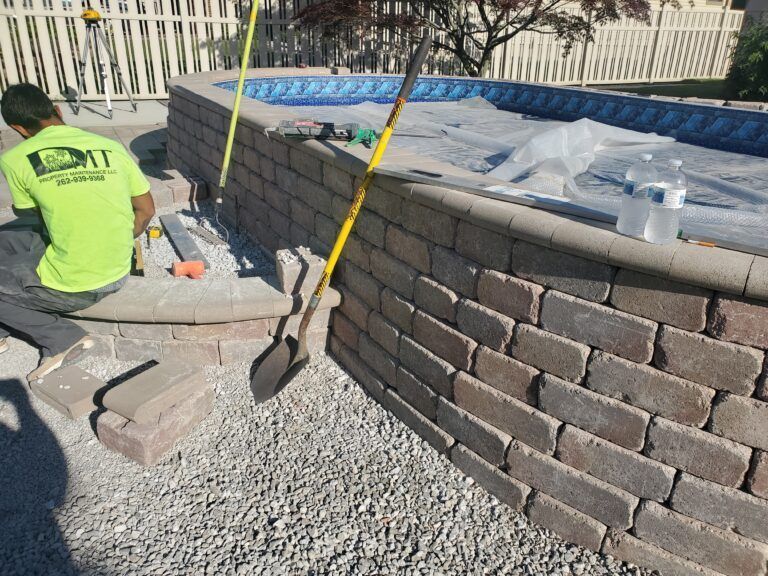 A worker in a neon green shirt builds a stone retaining wall around an above-ground swimming pool.