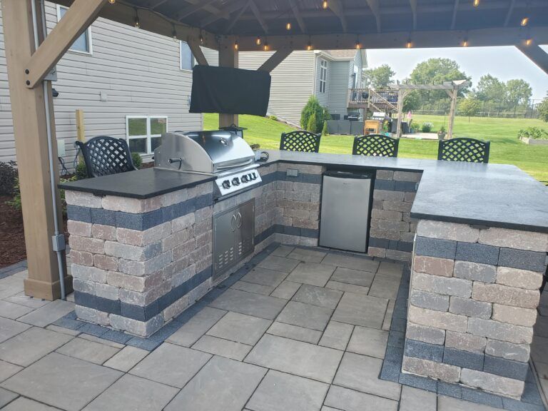 An outdoor stone kitchen with a grill, mini-fridge, and counter seating under a wooden pergola on a paved patio.