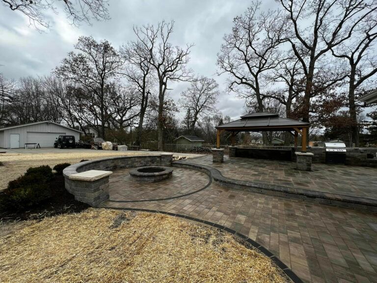 A backyard patio features a stone fire pit, a raised semicircular retaining wall, and a pavilion under cloudy skies.