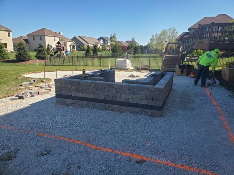 A worker in a neon jacket builds a stone patio wall in a sunny residential backyard.