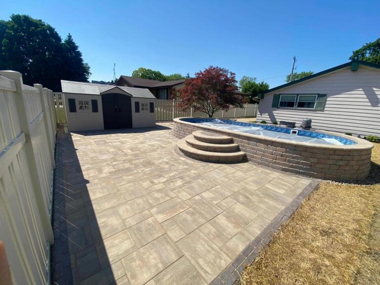 A paved backyard patio featuring a tan rectangular stone surface, a small dark shed, and an above-ground pool with stone steps.