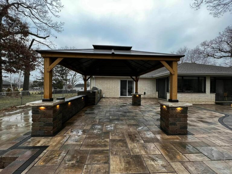 A backyard patio with a wooden gazebo covering a stone bar and dining area, set against a house on an overcast day.
