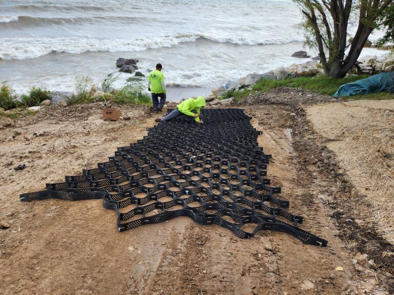 Two workers in neon vests install a black honeycomb-shaped geogrid erosion control mat on a slope near a wavy shoreline.