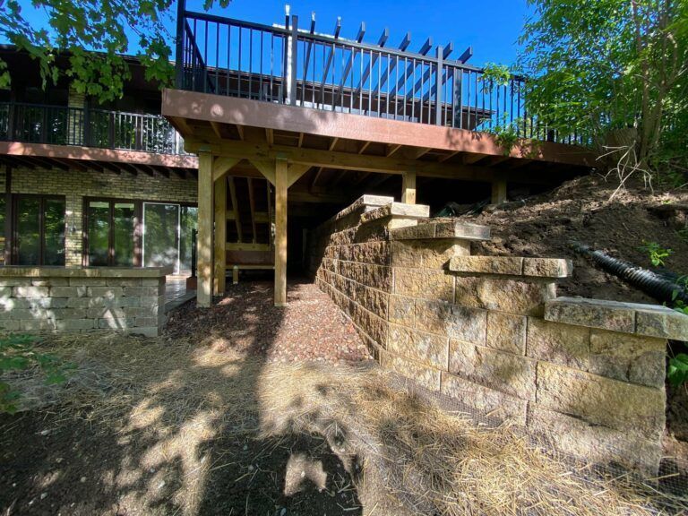 A wooden deck overlooks a stone retaining wall and a path leading beneath the structure on a sunny day.