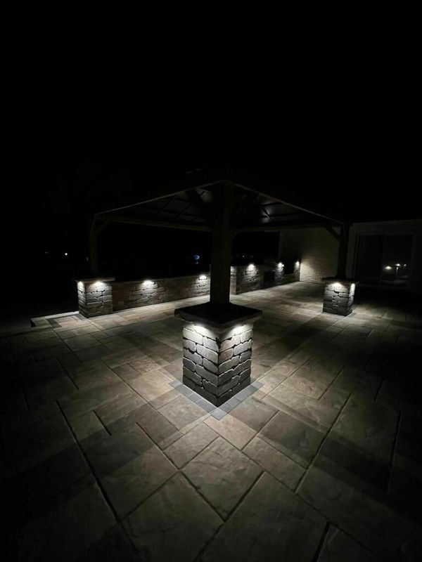 A stone patio at night, illuminated by recessed lights on square stone columns and a low wall under a dark pergola.