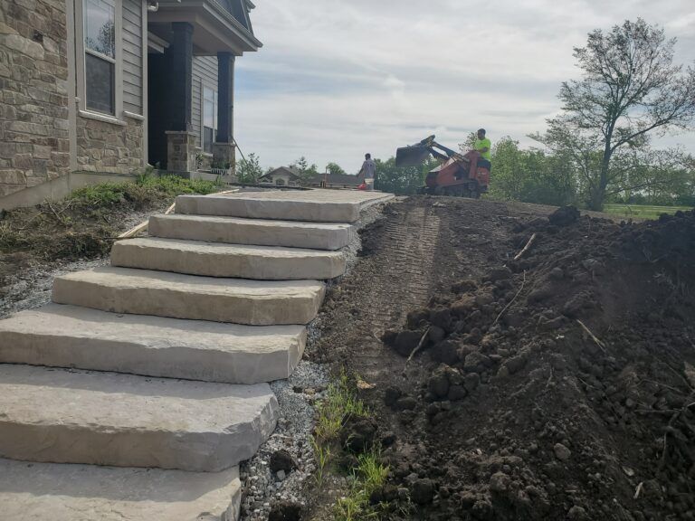 New stone steps are being installed next to a house under construction while a worker operates a skid steer nearby.