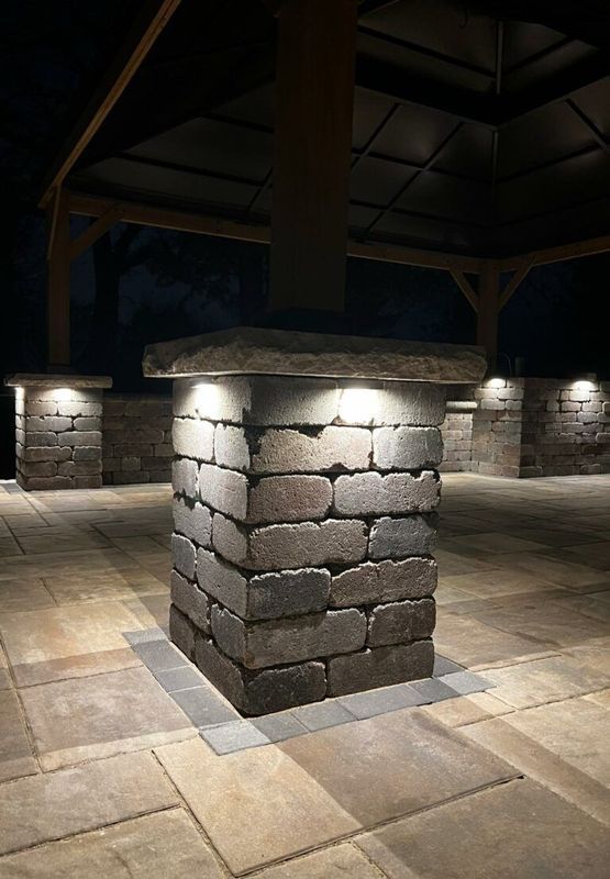 Outdoor stone pillar base with integrated accent lighting under a pergola at night, set on a large stone-paved patio.