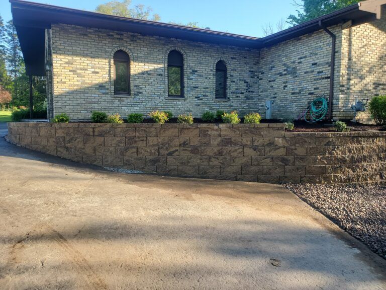 A tan stone house features three arched windows above a curved, tiered retaining wall in a paved driveway setting.