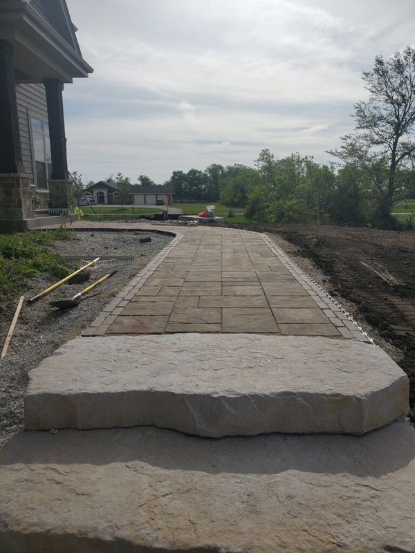A stone walkway leads from a large natural stone step toward a house under construction on a sunny day.