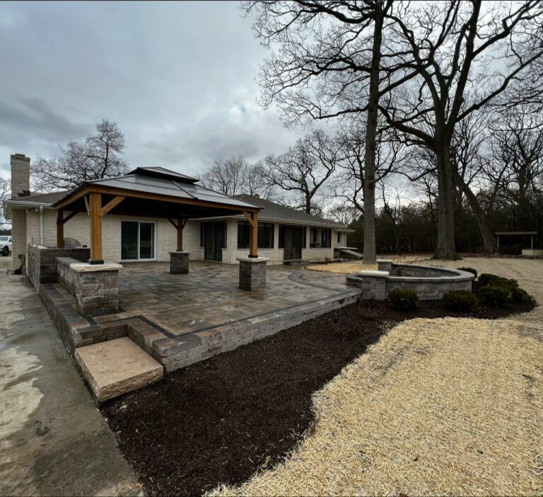 A stone patio with a gazebo, retaining walls, and mulch landscaping in front of a house on a cloudy day.