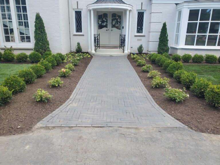 A gray stone paver walkway leads to a white front door, flanked by mulched beds with small shrubs and two tall evergreens.