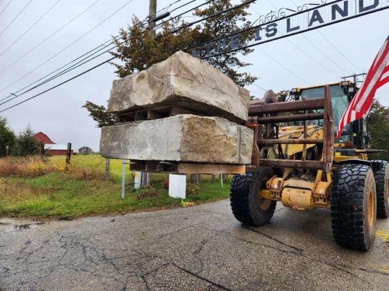 A yellow front-end loader lifts two large, stacked stone blocks on a wooden pallet along a paved road.