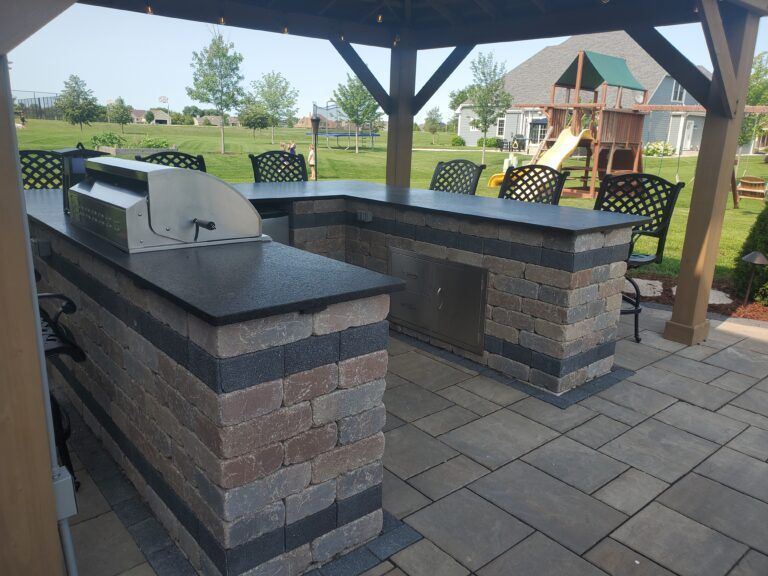 An L-shaped stone outdoor bar with a built-in grill and chairs, situated on a paver patio under a wooden gazebo.