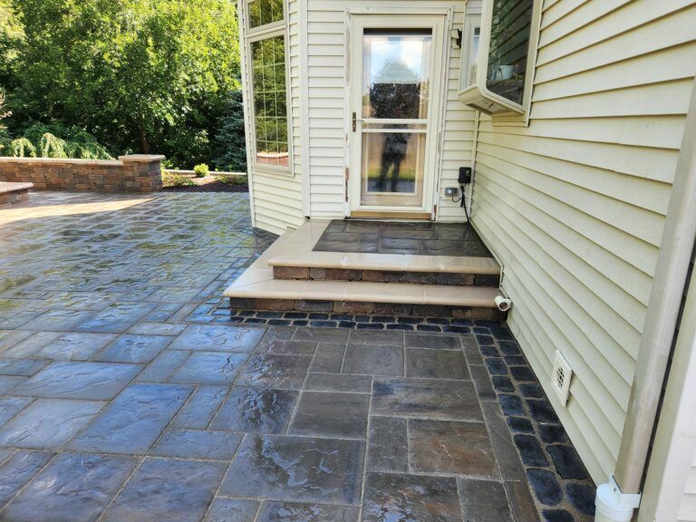 A home's exterior features a stone paver patio leading to two steps and a storm door, bordered by a dark stone accent edge.