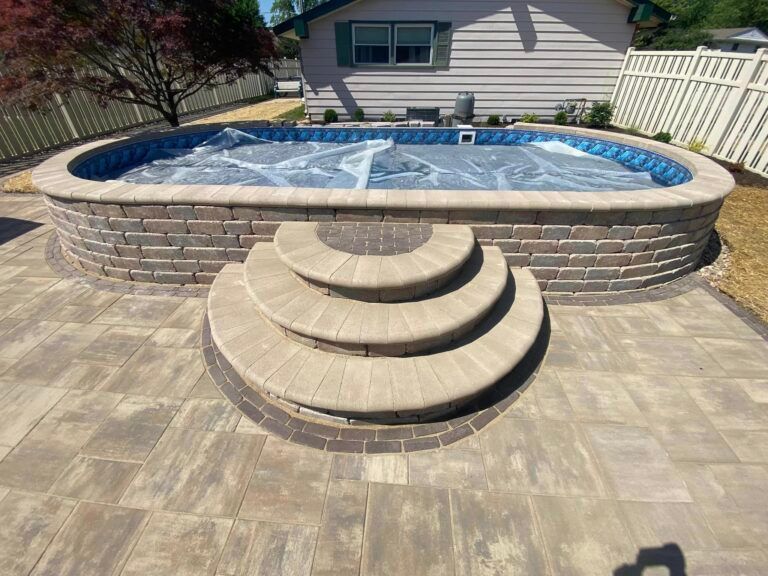 An oval, above-ground swimming pool with a stone wall and built-in stone steps on a patio in a backyard.