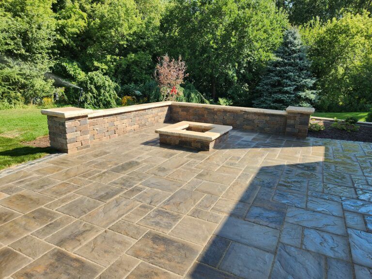 A stone patio with a built-in square fire pit and a low retaining wall, set against a background of green trees.