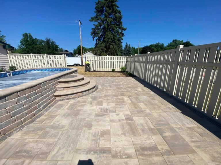 A tan paver patio area with a built-in swimming pool and semi-circular stone steps, enclosed by a white vinyl fence.