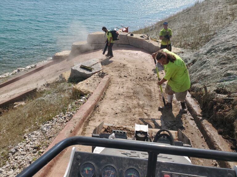 Workers in bright green shirts construct a dirt path along a shoreline, using a leaf blower and shovel near a vehicle.