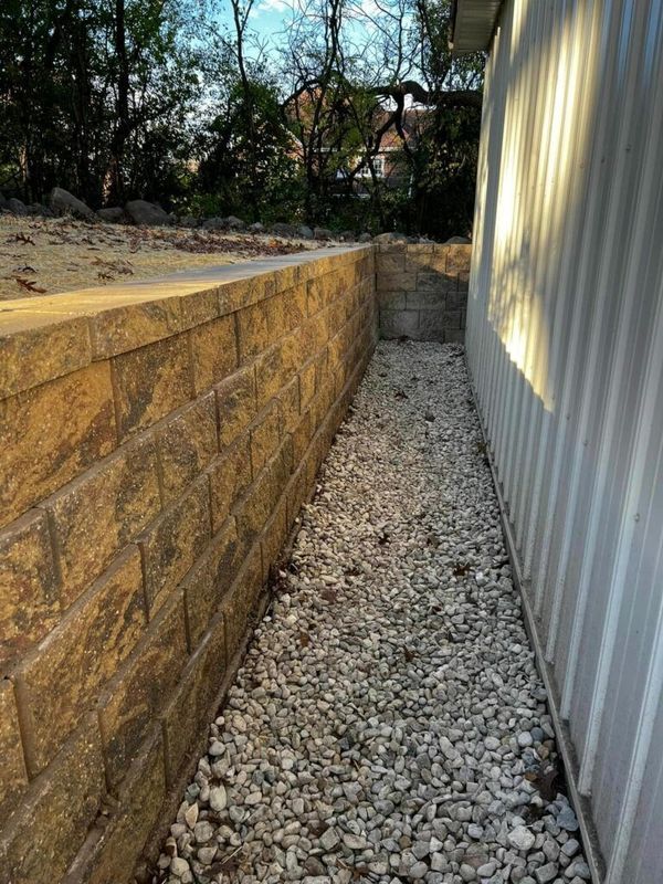 A stone retaining wall sits parallel to a metal wall with a gravel path in between, outdoors under bright sunlight.