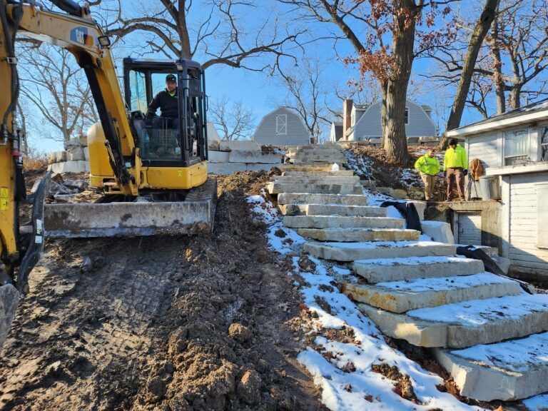 A yellow excavator sits beside a newly constructed stone staircase on a snowy, sloped lot near a house.