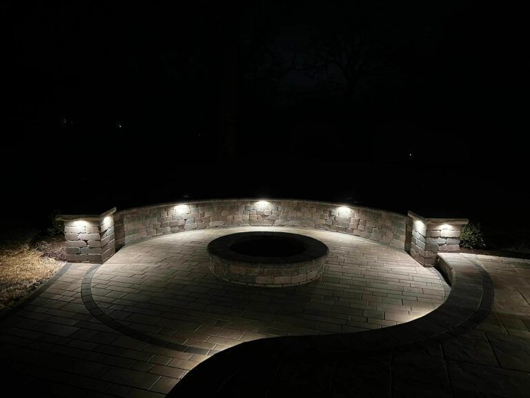 A lit outdoor fire pit area at night featuring a curved stone bench with integrated lighting on a paved patio.