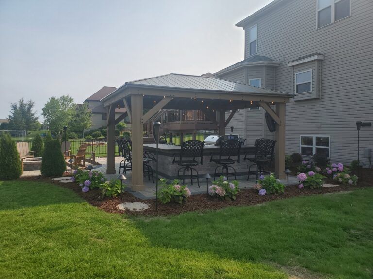 A wooden gazebo covering a patio bar with chairs, set in a backyard near a house with landscaping and a grassy lawn.