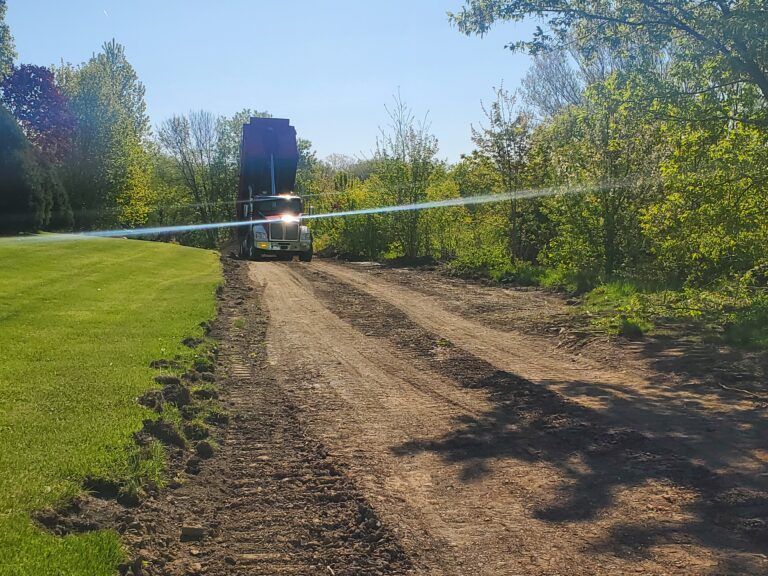 A large dump truck moves along a dirt construction path beside a grassy field surrounded by trees under a clear blue sky.