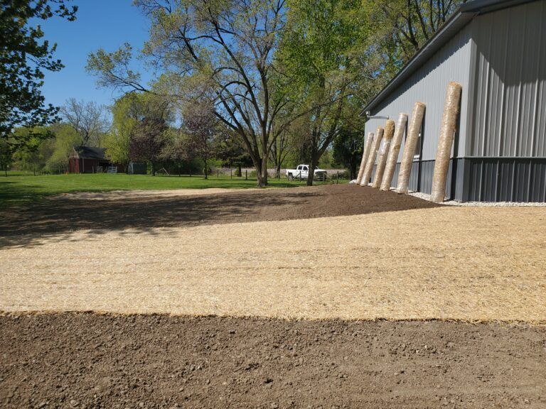 A recently seeded yard with straw matting and bare soil next to a large grey metal building under a clear blue sky.