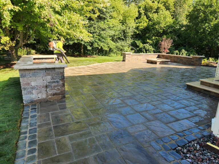 A person uses a leaf blower on a landscaped patio with stone pavers, retaining walls, and a fire pit near a grassy area.