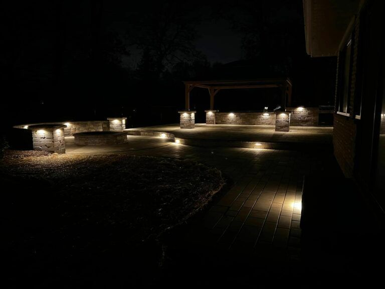 A stone patio at night, illuminated by warm landscape lighting on low walls, steps, and a pergola-covered outdoor kitchen.