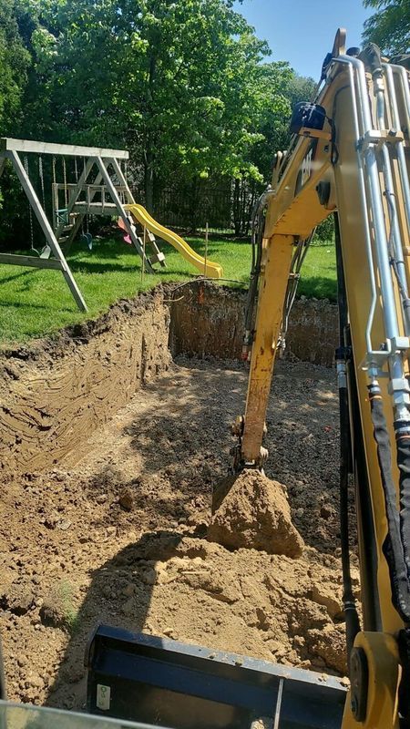Excavator arm digging a large hole in a backyard next to a wooden playset with a yellow slide.
