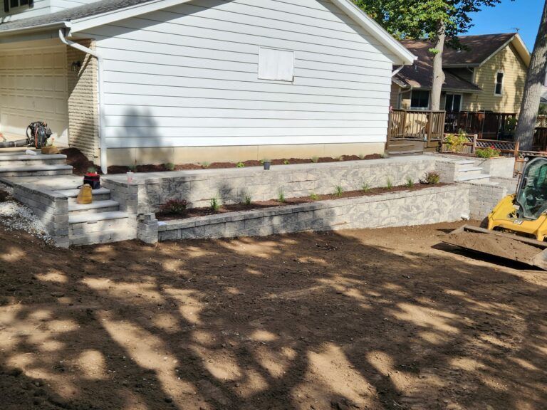 New light-colored stone retaining walls and stairs in front of a white house with a yellow excavator nearby.