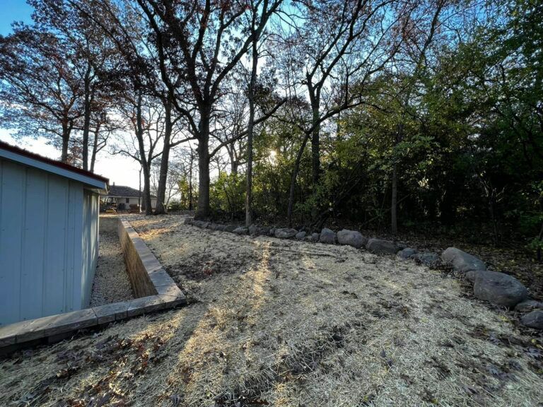 A retaining wall borders a sloped yard covered in light-colored mulch, with a house corner and mature trees in the background.