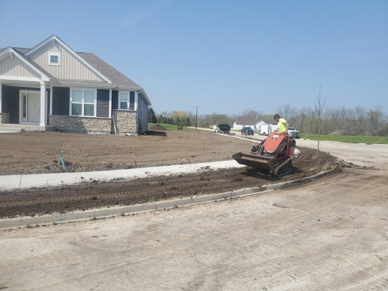 A construction worker operates a small tracked loader to grade dirt along a residential sidewalk near a new home.