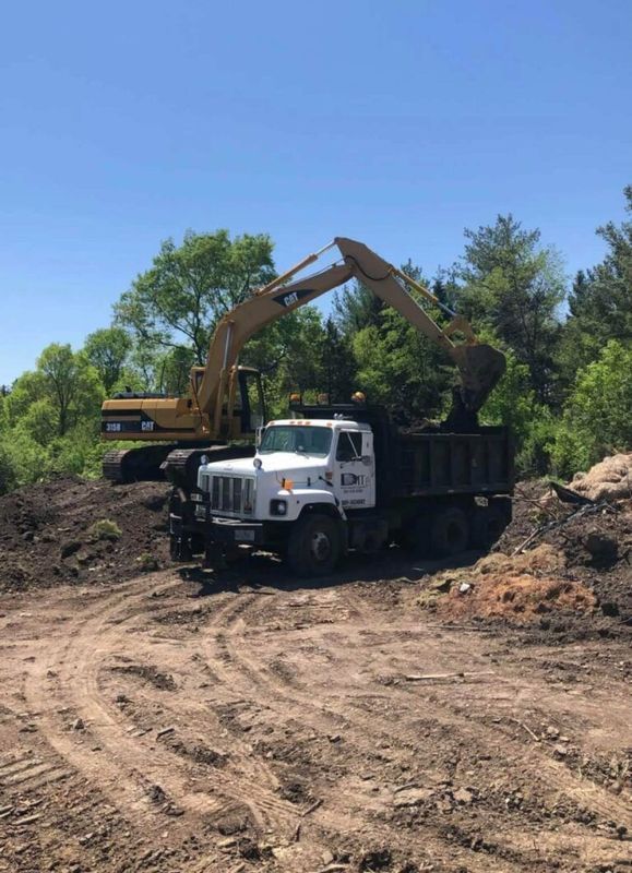 A tan excavator loading soil into the back of a white dump truck at an outdoor construction site.