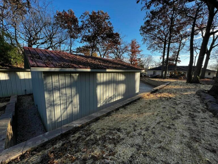 A pale green shed with a red metal roof sits in a grassy yard, bordered by a low concrete retaining wall under trees.
