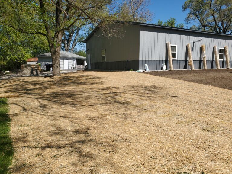 A gray metal building with wooden support beams next to a field of dormant grass under a bright blue sky.
